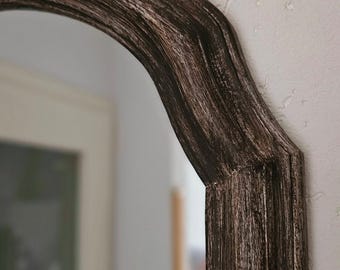 Pode incluir: Um close-up de uma moldura de espelho de madeira preta com acabamento envelhecido. A moldura tem o topo arredondado e a parte inferior reta.