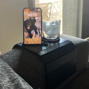 Puede incluir: Una bandeja negra para el brazo del sofá con un teléfono inteligente que muestra una imagen de un perro, un vaso de agua con hielo y un cable de carga. La hora en el teléfono es 10:15. La bandeja está sobre un sofá gris.