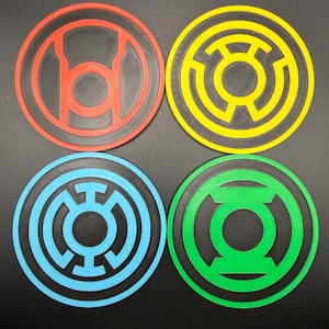 May include: Four colorful round coasters featuring superhero symbols. The coasters are red, yellow, blue, and green, each with a unique symbol design. The coasters are made of a textured material and are set against a black background.