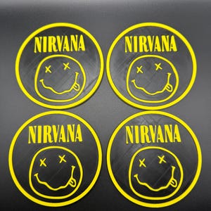May include: Four black coasters with a yellow Nirvana logo. Each coaster features the band's name and a smiley face with crossed-out eyes and a protruding tongue. The coasters are round and have a textured surface.