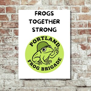 May include: A white poster with the text "FROGS TOGETHER STRONG" in black. Below is a green circle with a cartoon frog wearing a helmet and giving a peace sign. The circle reads "PORTLAND FROG BRIGADE" in a circular font.