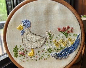 May include: Embroidered artwork featuring a duck wearing a blue bonnet, walking with three yellow ducklings. The scene includes green grass, pink and red flowers, and a blue stream. The embroidery is set in a round wooden hoop.