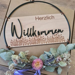 以下が含まれることがあります： ドイツ語の「Willkommen」の黒い文字と「Herzlich」の茶色の文字が入った木製のウェルカムサイン。サインはドライフラワーと紫のリボンで飾られており、濃い緑色の金属リングで囲まれています。
