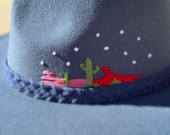 Pode incluir: Um chapéu de cowboy de feltro azul com uma banda trançada e cactos e flores bordadas. O chapéu tem costuras brancas ao redor da borda.