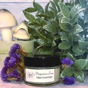 May include: A small glass jar with a black lid containing a light green balm. The jar has a white label with the text "Magnesium Lane Tallow Facial Balm". The jar is sitting on a wooden surface with green plants and purple flowers.
