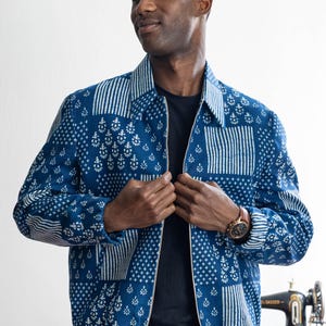 Peut inclure: Veste bleue à motifs avec fermeture éclair. La veste présente un motif patchwork avec des motifs géométriques blancs et bleus. La personne porte un t-shirt noir et une montre avec un bracelet en cuir marron.