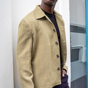 Könnte beinhalten: Eine hell olivgrüne Jacke mit Kragen und Knopfverschluss. Die Jacke wird über einem schwarzen Hemd getragen und mit einer dunkelblauen Hose kombiniert. Die Jacke hat ein strukturiertes Aussehen und wird von einer Person getragen.