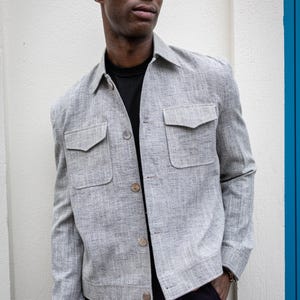 Peut inclure: Veste gris clair avec deux poches avant et des boutons. La veste est portée sur un t-shirt noir. Le modèle porte un pantalon bleu foncé. La veste a un aspect texturé.