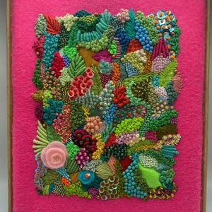 Op de afbeelding: Een ingelijst textielkunstwerk met een levendig, gestructureerd ontwerp. Het kunstwerk toont geborduurde elementen in tinten groen, blauw, rood en roze, tegen een felroze achtergrond. De lijst is gemaakt van lichtgekleurd hout.