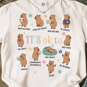 May include: Cream-colored t-shirt featuring a graphic of capybaras in various poses with phrases like "Make Mistakes" and "Be Yourself." The words "It's ok to" are in the center. The shirt has a crew neck and short sleeves.