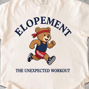 May include: Off-white t-shirt featuring the word "ELOPEMENT" in navy blue, arched across the top. A cartoon bear in a red headband, navy vest, and red shorts is running. The text "THE UNEXPECTED WORKOUT" is below the bear.