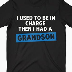 May include: Black t-shirt with white text that reads "I USED TO BE IN CHARGE THEN I HAD A GRANDSON." The word "GRANDSON" is in a blue rectangle. The shirt is a short-sleeved crew neck.