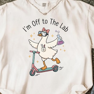Könnte beinhalten: Weißes T-Shirt mit einer Grafik einer Gans mit Brille und Schleife, die auf einem rosa und türkisfarbenen Roller fährt und einen Becher hält. Der Text lautet "I'm Off to The Lab".