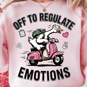 May include: Pink sweatshirt with the text "OFF TO REGULATE EMOTIONS". The graphic features a white goose riding a pink scooter with a backpack, a heart, and a peace sign.
