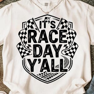 Op de afbeelding: Wit t-shirt met een zwarte afbeelding met de tekst "It's Race Day Y'all". Het ontwerp bevat geblokte vlaggen en bandensporen, wat een racethema oproept. Het shirt is gemaakt van een zacht materiaal.