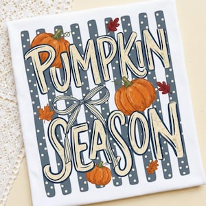 May include: A white t-shirt featuring a graphic print with the text PUMPKIN SEASON in cream letters. The design includes three orange pumpkins, a grey and white striped bow, and small red and orange autumn leaves over a grey polka dot background.