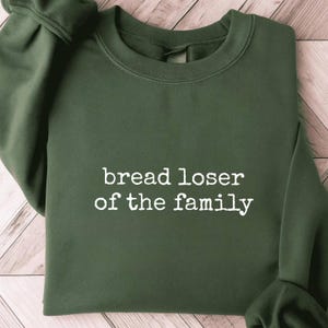 May include: An olive green sweatshirt with the phrase "bread loser of the family" printed in white. The crew neck sweatshirt is folded on a light wood grain surface.