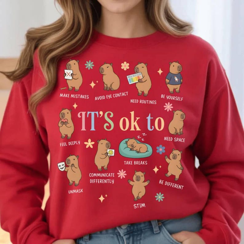 May include: Red sweatshirt with the phrase "It's ok to" and illustrations of capybaras. The sweatshirt features various phrases like "Make Mistakes," "Avoid Eye Contact," and "Be Yourself."