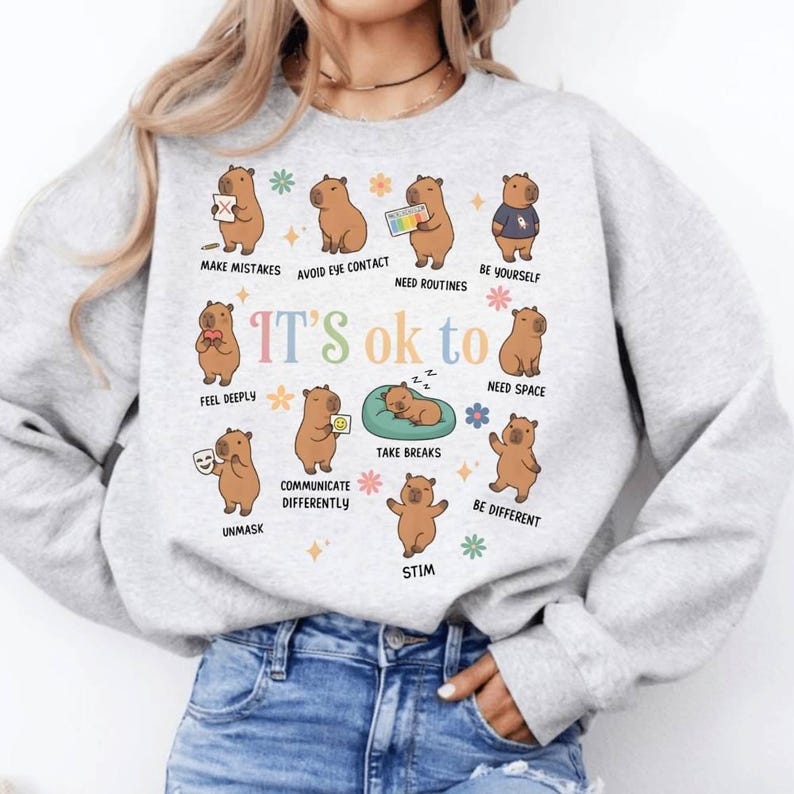 May include: A light gray sweatshirt featuring cartoon capybaras with various phrases like "Make Mistakes" and "Be Yourself." The words "It's ok to" are in the center. The design includes colorful floral accents and stars.