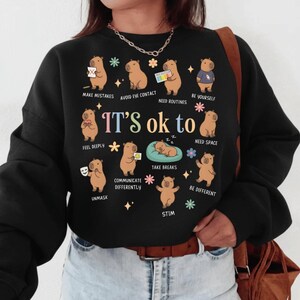 May include: Black sweatshirt with a graphic featuring capybaras and the text "IT'S ok to." The capybaras are depicted in various poses with phrases like "Make Mistakes," "Be Yourself," and "Take Breaks." The design includes colorful floral accents.