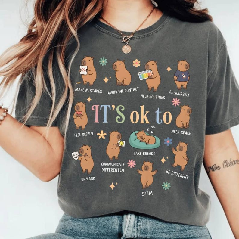 May include: Dark gray t-shirt with the phrase "IT'S ok to" and illustrations of capybaras. The shirt features various phrases like "Make Mistakes", "Avoid Eye Contact", and "Be Yourself". The design includes colorful flowers and stars.