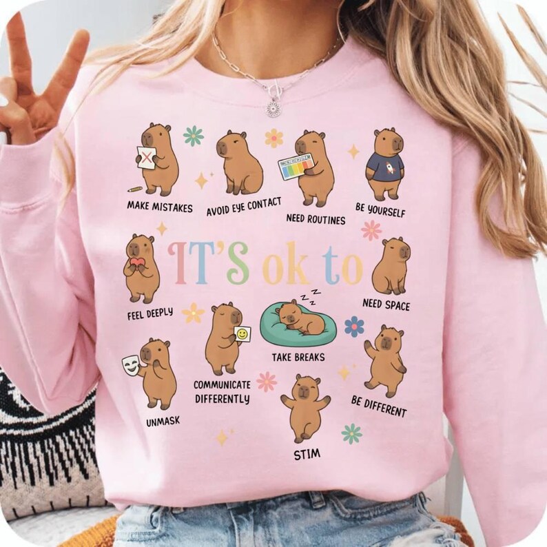 May include: Pink sweatshirt with the phrase "IT'S ok to" and illustrations of capybaras. Each capybara represents a different concept, such as "Make Mistakes," "Be Yourself," and "Take Breaks." The sweatshirt is a soft pink color.