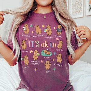 May include: A purple t-shirt with the phrase "It's ok to" and various affirmations. The shirt features cartoon capybaras illustrating concepts like "Make Mistakes" and "Be Yourself". The design includes colorful floral accents.