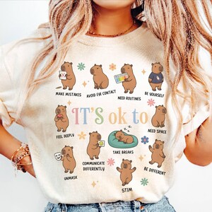 May include: Cream-colored t-shirt with a colorful graphic of capybaras illustrating various emotional states. The text "IT'S ok to" is above the capybaras, with phrases like "Make Mistakes" and "Be Yourself" below them. The shirt has short sleeves.