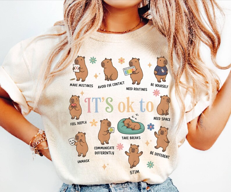 May include: Cream-colored t-shirt with a colorful graphic of capybaras illustrating various emotional states. The text "IT'S ok to" is above the capybaras, with phrases like "Make Mistakes" and "Be Yourself" below them. The shirt has short sleeves.