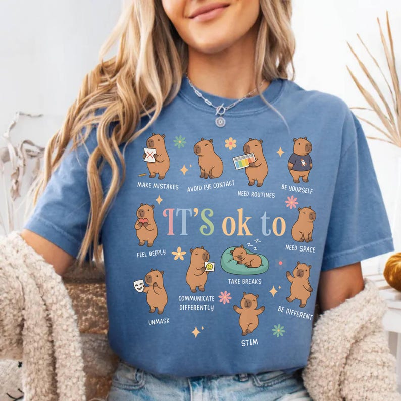 May include: Blue t-shirt with a graphic featuring capybaras and the text "IT'S ok to". The capybaras are depicted in various poses with phrases like "Make Mistakes", "Avoid Eye Contact", and "Be Yourself".