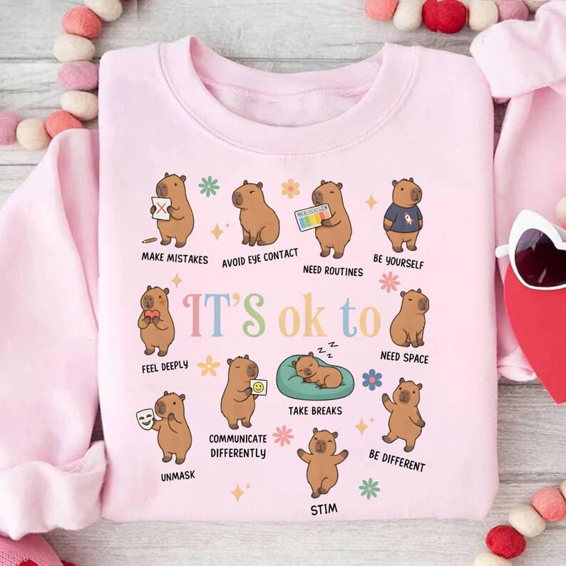 May include: A light pink sweatshirt with a colorful graphic featuring capybaras and the text "IT'S ok to." Each capybara illustrates a different concept, such as "Make Mistakes" and "Be Yourself." The design includes flowers and stars.
