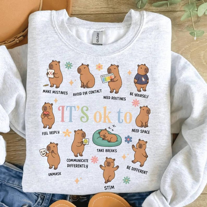 May include: Light gray sweatshirt with a colorful graphic of cartoon capybaras. Each capybara is depicted with a different action, and the text "IT'S ok to" is in the center. The sweatshirt has a crew neck and long sleeves.