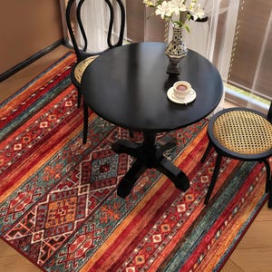 May include: A round black table with two black chairs and a colorful rug. The rug features a geometric pattern with red, orange, and teal stripes. A cup of coffee sits on the table, next to a vase of white flowers.