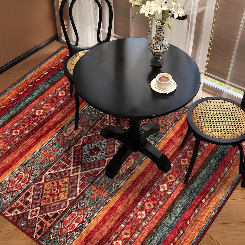 May include: A round black table with two black chairs and a colorful rug. The rug features a geometric pattern with red, orange, and teal stripes. A cup of coffee sits on the table, next to a vase of white flowers.