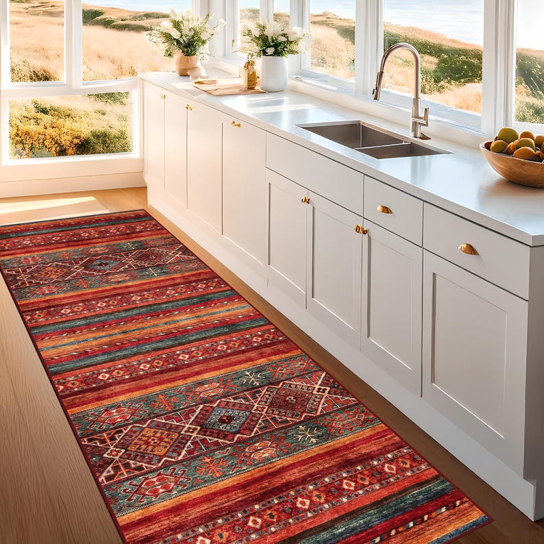 May include: A long, rectangular rug with a vibrant, geometric pattern. The rug features a mix of red, orange, teal, and brown stripes and diamond shapes. It is placed on a wooden floor in front of a white kitchen cabinet.