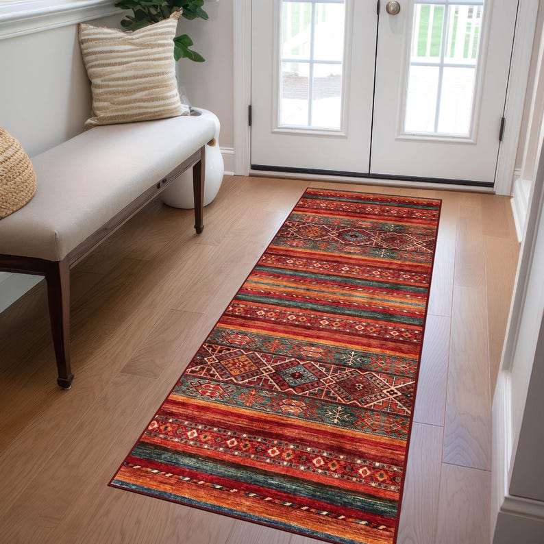 May include: A rectangular runner rug with a vibrant, geometric pattern. The rug features a mix of red, orange, teal, and brown hues, with horizontal stripes and diamond-shaped motifs. It is placed on a light-colored wooden floor, near a bench and a white door.