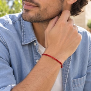 May include: A thin, vibrant red cord bracelet worn on the wrist. The bracelet consists of a simple, smooth, and minimalist red string that wraps around the wrist, providing a subtle and understated accessory that complements the casual blue shirt.