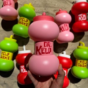 Pode incluir: Uma coleção de garrafas coloridas em rosa, verde e vermelho. Cada garrafa apresenta o texto "DR. KELD" e "KELD SHAKE" em negrito vermelho. As garrafas têm um design único e bulboso.
