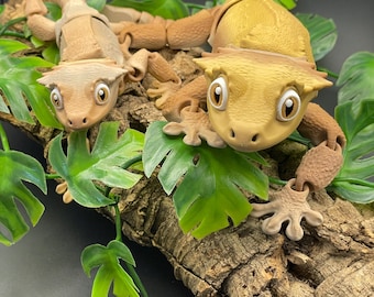 May include: Two brown and tan gecko figurines with large eyes and textured skin, posed on a piece of bark with green leaves.