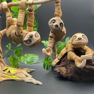 May include: Three brown sloth figurines with articulated limbs, posed on a tree branch and a log. The sloths have realistic features, including large eyes and a furry texture.