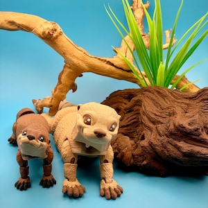 May include: Two articulated otter figurines in tan and brown, posed on a light blue surface. The otters are positioned near a piece of driftwood and green artificial grass. The background is a solid light blue.