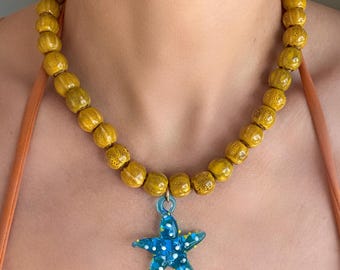 May include: A necklace featuring large, yellow beads with a blue starfish pendant. The starfish is decorated with white and yellow dots and hangs from a blue loop. The necklace is worn around the neck.