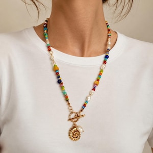 Op de afbeelding: Een kleurrijke kralenketting met een goudkleurige hanger. De ketting heeft een verscheidenheid aan kralen in regenboogkleuren, afgewisseld met witte parels. De hanger is een gedetailleerd, cirkelvormig ontwerp met een kleine parel.