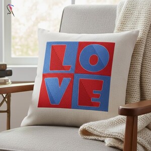 May include: A square throw pillow with the word "LOVE" in red and blue block letters. The letters are stitched onto a cream-colored fabric. The pillow is on a gray chair with a cream-colored blanket draped over it.
