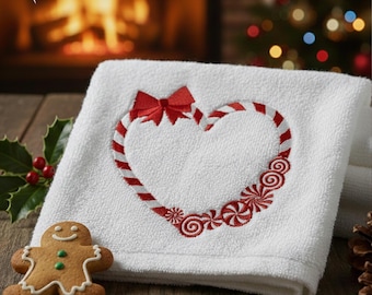 Könnte beinhalten: Weißes Handtuch mit einem Zuckerstangen-Herz-Design und einer roten Schleife. Das Herz ist mit rot-weißen Streifen umrandet und von gestickten Pfefferminzbonbons umgeben. Ein Lebkuchenmann-Keks liegt auf der Holzoberfläche.