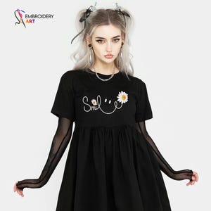 May include: Black short-sleeved dress with a white daisy and the word "Smile" in script. The dress has a gathered skirt and is paired with sheer black arm sleeves. The image also shows a logo for "Embroidery Art".