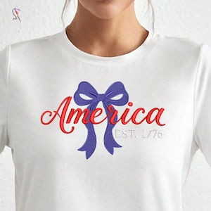 Puede incluir: Camiseta blanca con la palabra "America" en rojo y un diseño de lazo azul. El texto "EST. 1776" está debajo de la palabra. El diseño está bordado en la camiseta.