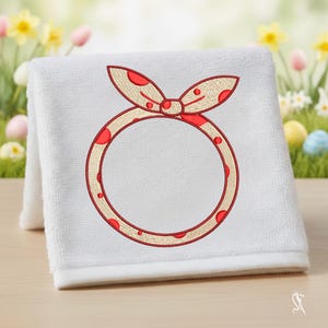 Op de afbeelding: Wit opgevouwen handdoek met een geborduurd ontwerp. Het ontwerp heeft een rood en beige stippen strik en een rode stippen cirkel. De handdoek is gemaakt van een zacht, absorberend materiaal, perfect voor badkamer of keuken.