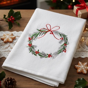 Könnte beinhalten: Weißes Küchentuch mit Weihnachts-Thema und gesticktem Kranzdesign. Der Kranz hat grüne Blätter, rote Beeren und eine rote Schleife. Das Handtuch ist gefaltet und liegt auf einer Holzoberfläche.