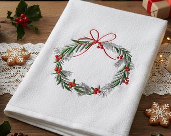 Könnte beinhalten: Weißes Küchentuch mit Weihnachts-Thema und gesticktem Kranzdesign. Der Kranz hat grüne Blätter, rote Beeren und eine rote Schleife. Das Handtuch ist gefaltet und liegt auf einer Holzoberfläche.